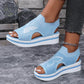 A pair of women's sky blue Nylira knitted wedge sandals with white butterfly embroidery, peep-toe design, and slingback strap, shown on feet against a textured stone-like background.