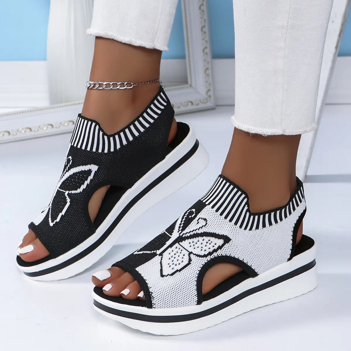 Women's black and white knit peep-toe wedge sandals with an embroidered butterfly motif and striped elastic collar, worn with white pants and an anklet.