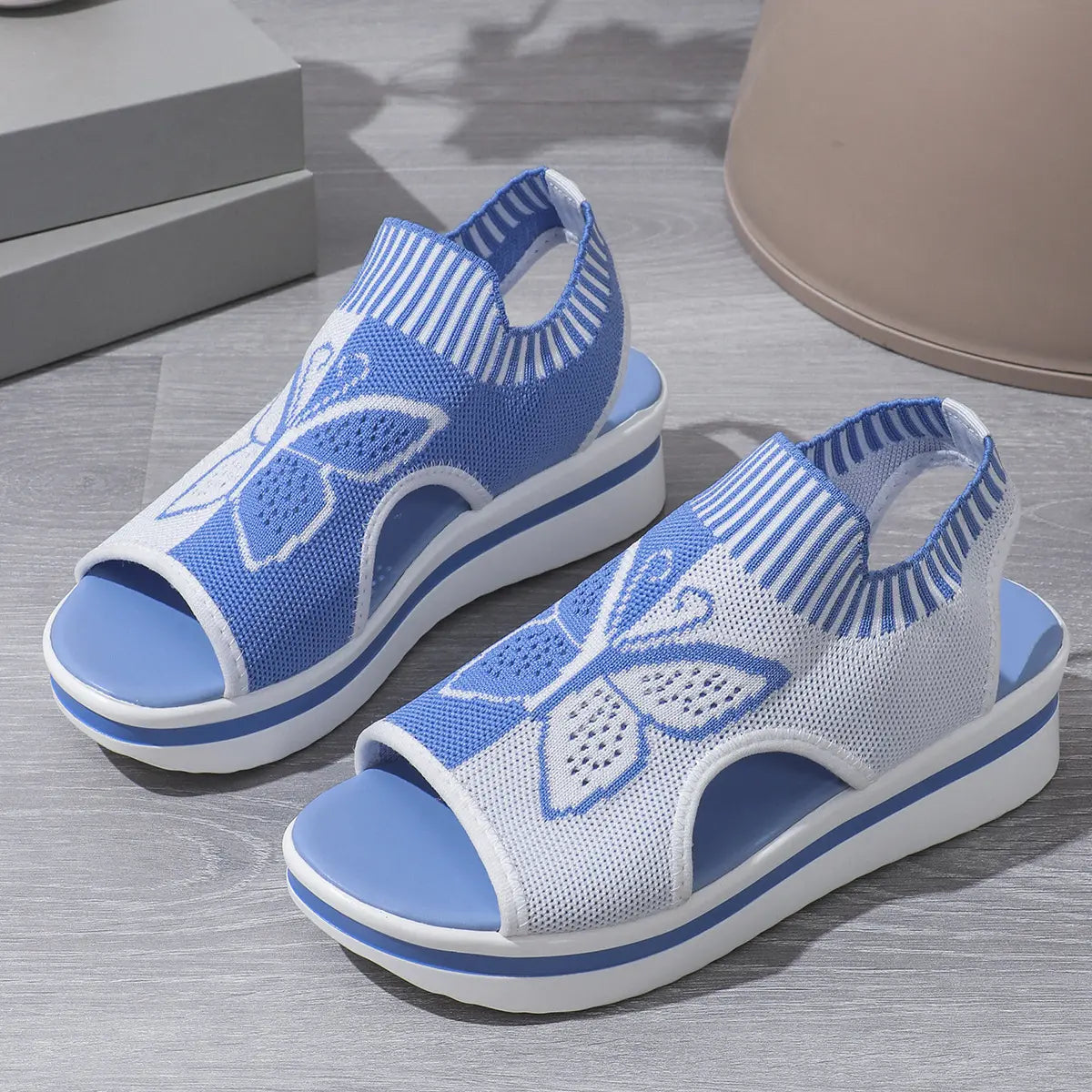 A pair of blue and white Nylira women's wedge sandals featuring a knitted butterfly design, peep-toe, and slingback strap. The layered wedge sole provides comfortable height.