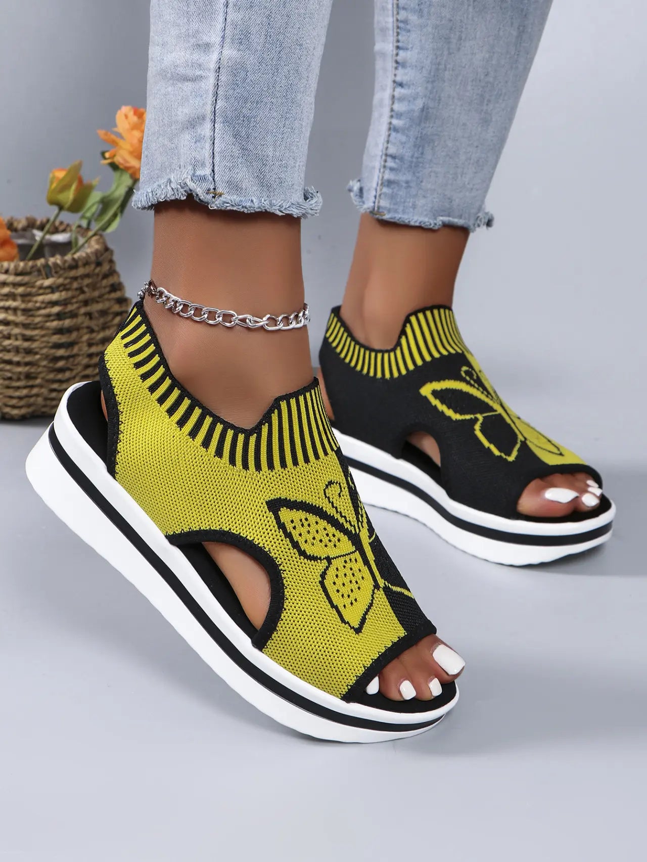 A pair of women's Nylira peep-toe wedge sandals in yellow and black, featuring a knitted butterfly design and a comfortable slingback strap. The sandals are worn with light-wash jeans, showcasing a stylish summer footwear option.