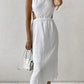 A white sleeveless midi dress with distinctive waist cut-outs and a crinkled bubble crepe texture is showcased on a model against a subtly textured wall.