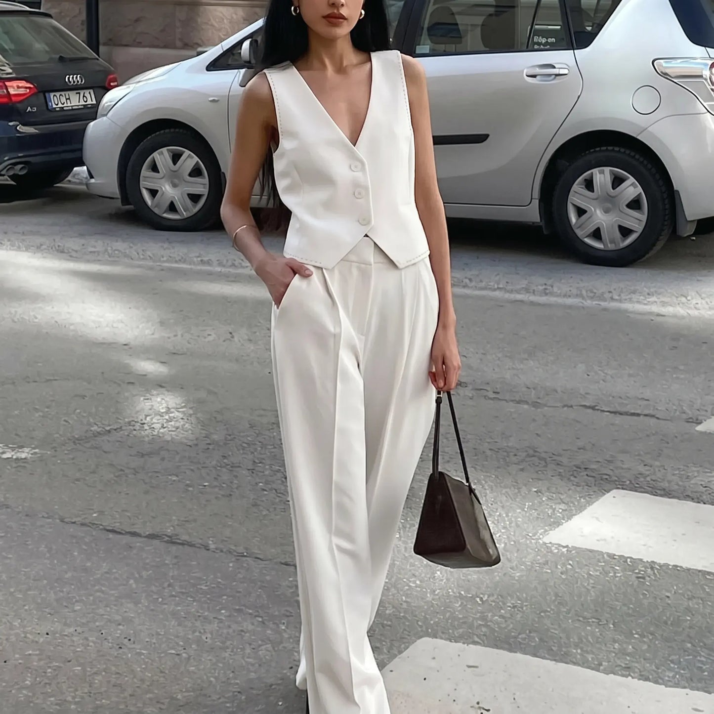 A woman models a pristine white sleeveless V-neck vest and flowing wide-leg trousers set, standing on a city street with a handbag.