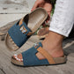 Ocean blue embellished slide sandals with tan accent straps and crystal butterfly detail, shown on a foot wearing white ripped jeans on a wooden deck.