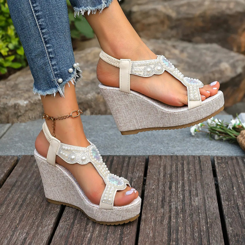 Close-up of off-white bohemian wedge sandals with intricate pearl and crystal embellishments on the T-strap, worn with distressed denim jeans. Features a high wedge heel covered in textured fabric and an elasticized ankle strap.