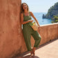 Woman wearing olive green cotton linen overalls with wide-leg fit and buckle straps, leaning against a wall in a coastal setting.
