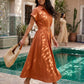 Woman walks confidently in an orange Zivro belted linen-blend A-line midi dress featuring short cuffed sleeves and an organic gold-tone buckle, carrying a straw bag in a beautiful riad setting.