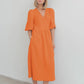 A woman models a vibrant orange cotton gauze midi wrap dress with short puff sleeves and functional side pockets, showcasing its flowing A-line silhouette. The dress is shown front-on against a clean white studio background.