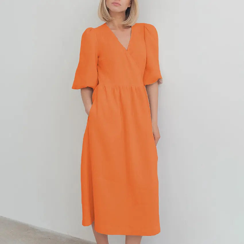A woman models a vibrant orange cotton gauze midi wrap dress with short puff sleeves and functional side pockets, showcasing its flowing A-line silhouette. The dress is shown front-on against a clean white studio background.
