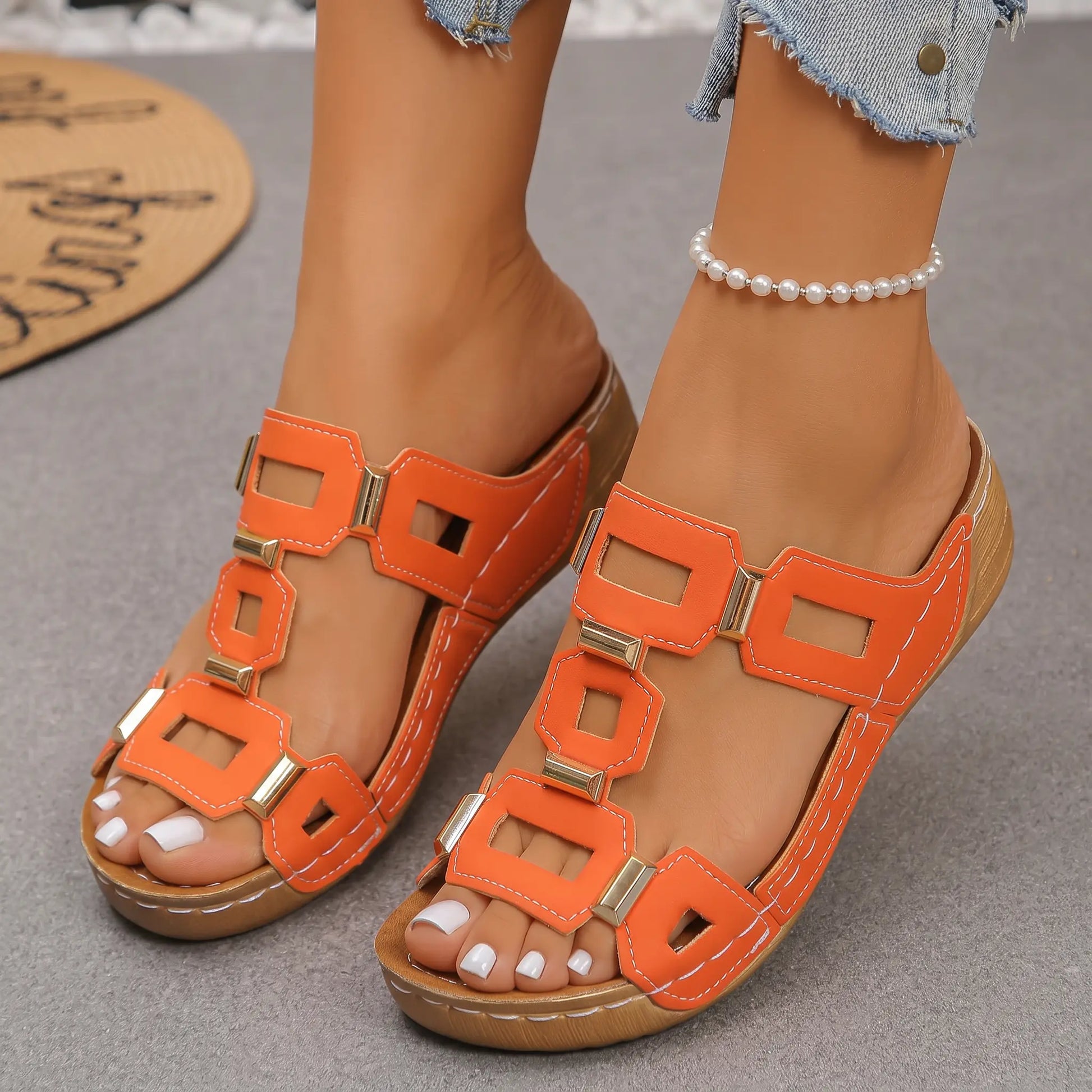 Vibrant orange platform wedge sandals featuring a geometric gold-tone embellishment design across the T-strap, shown on a model's foot with a pearl anklet and distressed denim.