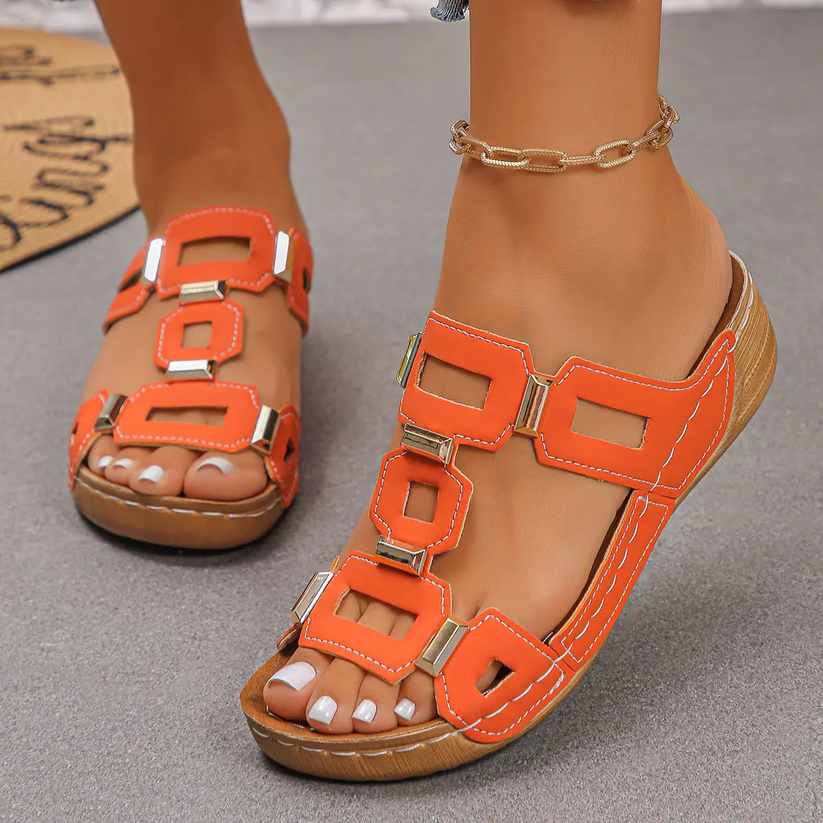 Orange platform wedge sandals with geometric gold-tone embellishments and white contrast stitching, worn on a foot with white pedicure and pearl anklet.