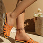 Vibrant orange woven flat slide sandals with an embellished block heel, styled on a model's foot with an anklet against a soft, sunlit backdrop.