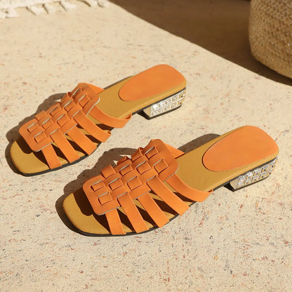 Vibrant orange woven flat slide sandals with an embellished block heel, displayed on a sunlit textured surface.