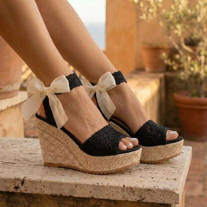 Oruzen Midnight Black Bohemian Espadrille Platform Wedges featuring a delicate lace peep-toe upper, natural woven espadrille platform, and elegant cream ribbon ankle ties, worn on feet outdoors.