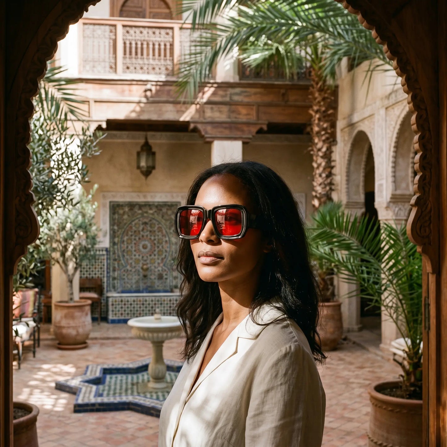 A woman wearing Kyzeth oversized square sunglasses with thick black frames and striking red lenses, standing in a sunlit Riad courtyard, embodying luxury and distinctive style.
