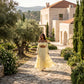 Model wearing a pale yellow bohemian off-shoulder tiered maxi set with long bell sleeves, walking on a sunlit stone path in a garden.