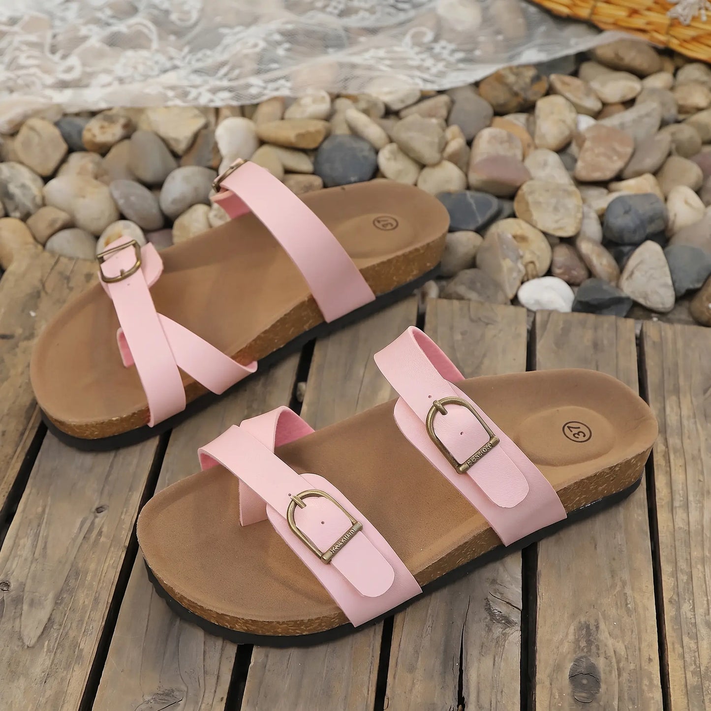 A pair of blush pink slip-on sandals with contoured cork footbeds and adjustable antique brass buckle straps, displayed on a rustic wooden surface with stones and lace.