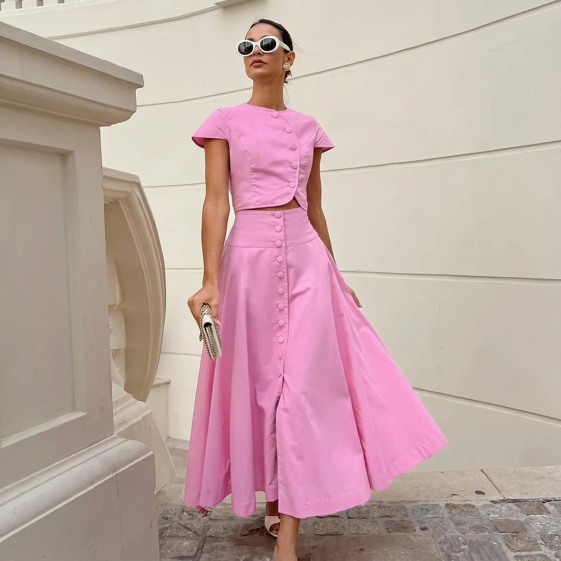 Vibrant pink cotton two-piece set featuring a cropped button-up top with cap sleeves and a flowing A-line midi skirt, modeled with sunglasses and a clutch.