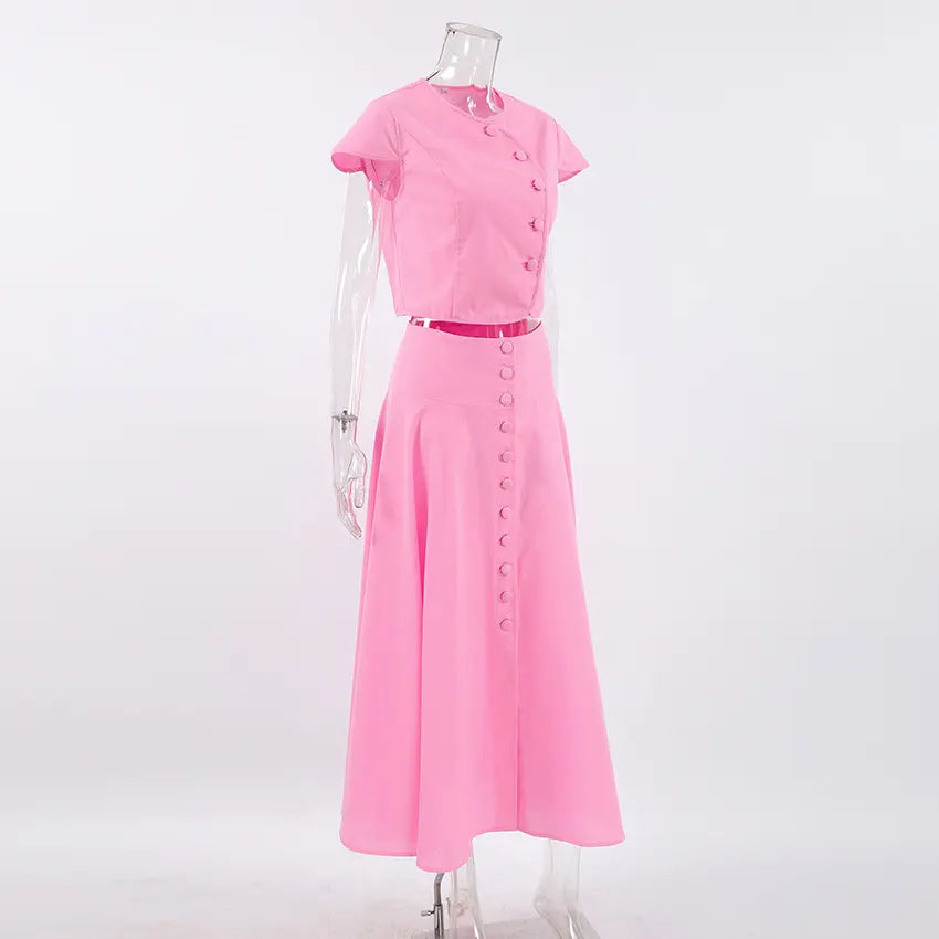 Vibrant pink cotton two-piece set on a mannequin, featuring a cropped button-up top with cap sleeves and a high-waisted, A-line midi skirt with a full button-down front, shown from a side angle.