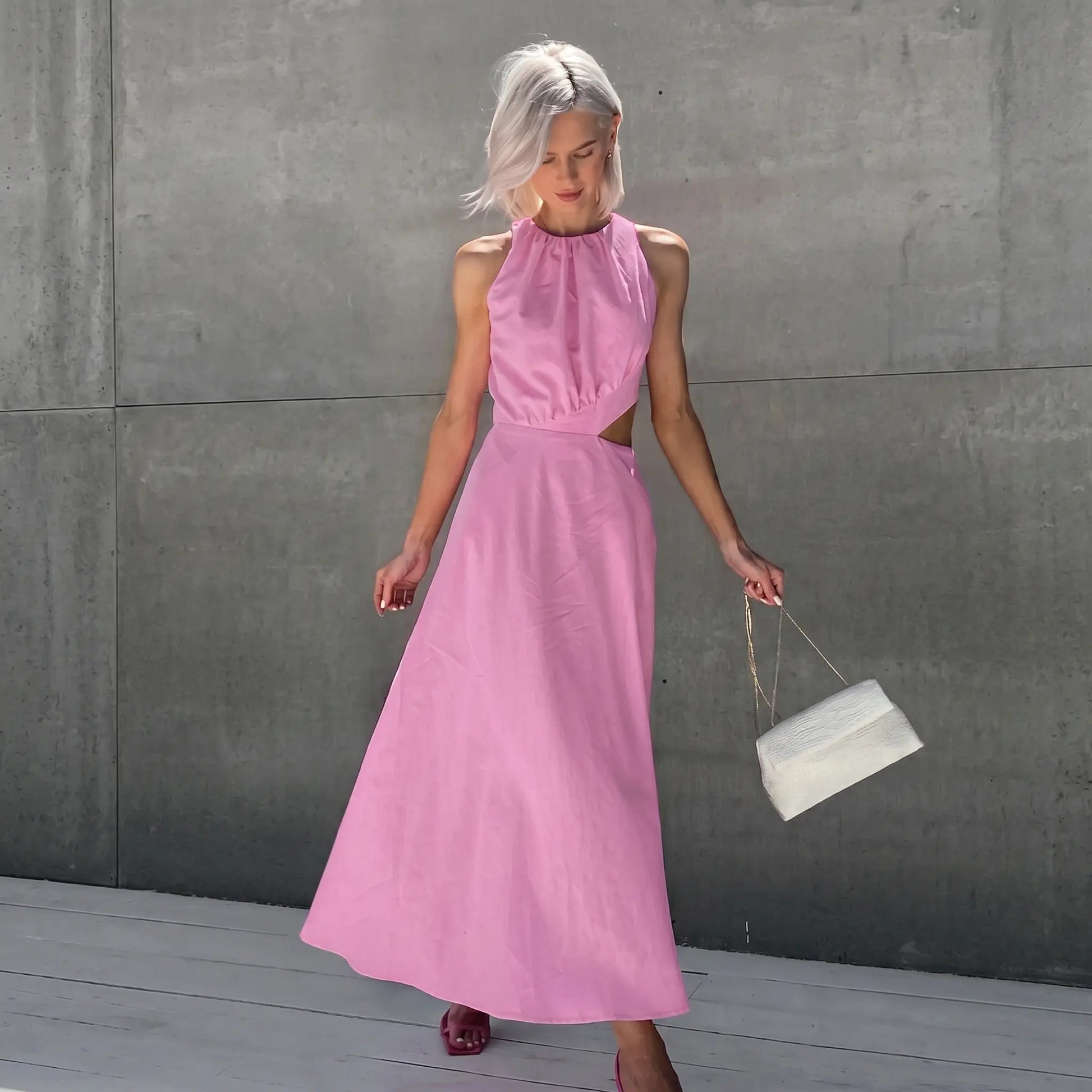 Elegant pink midi dress with gathered halter neckline and side waist cut-outs, worn by a model in a lifestyle setting. Features a flowy A-line skirt.