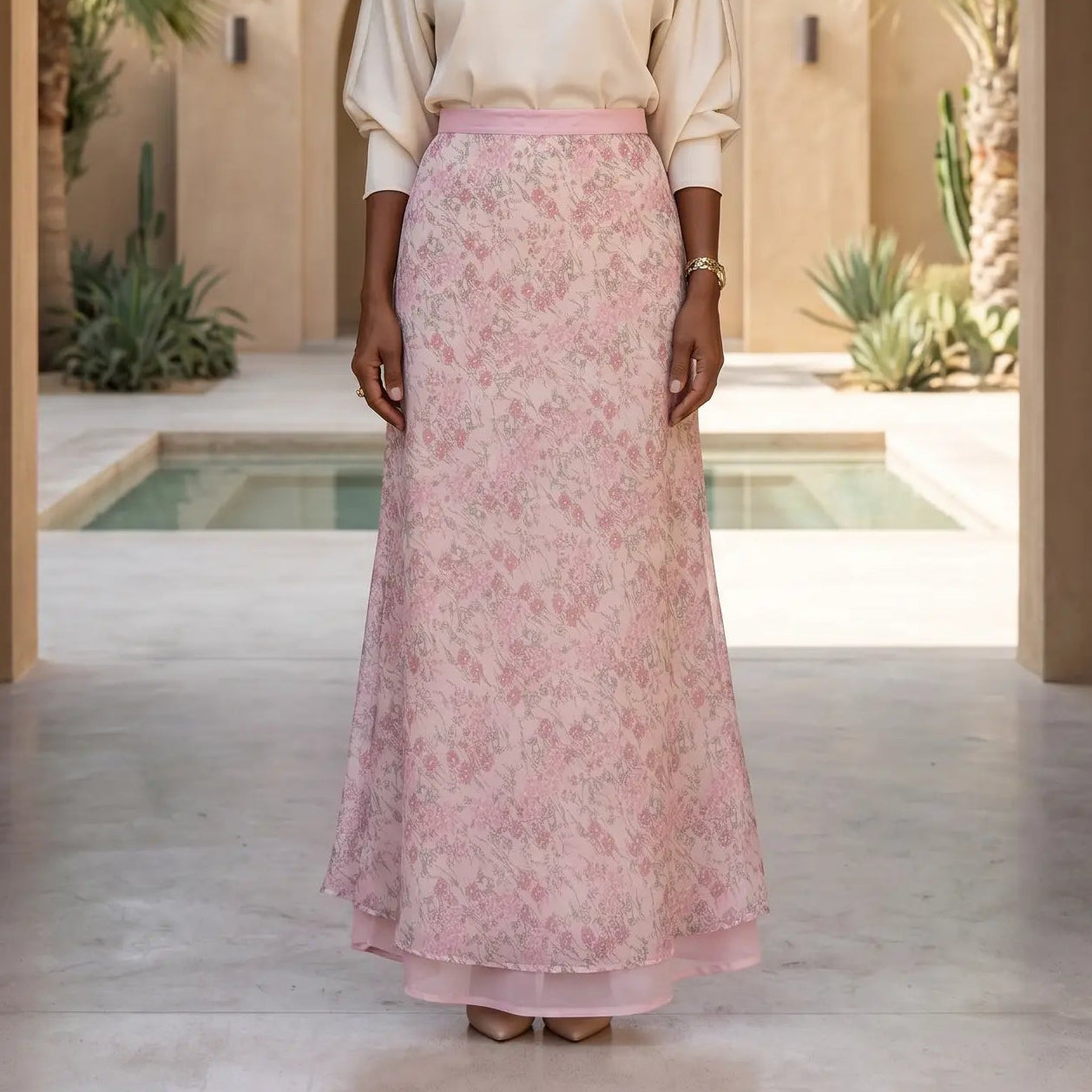 Elegant woman models a high-waisted pink ditsy floral chiffon maxi skirt with a fluid silhouette. Posed in an arched hall with desert plants.