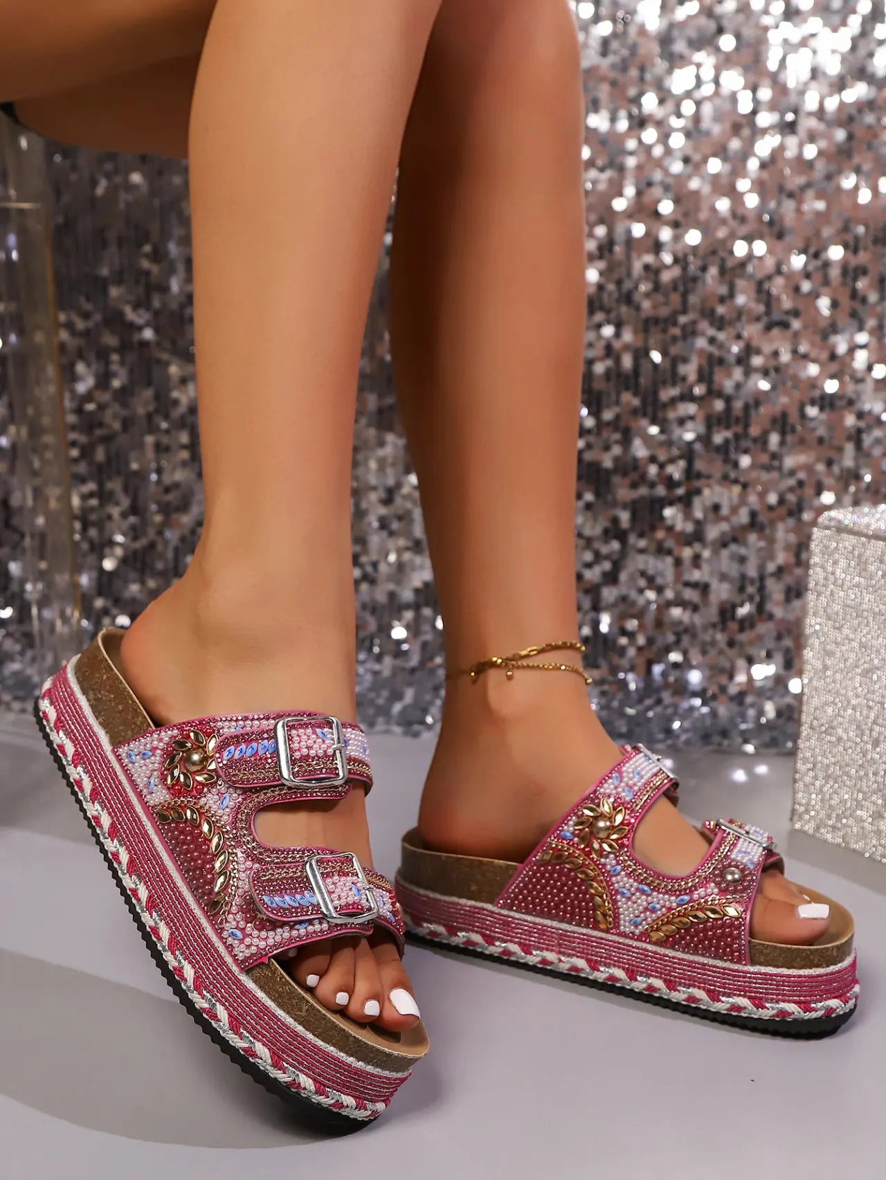 Rose pink platform slide sandals embellished with beads and sequins, featuring double adjustable buckle straps and a layered cork-effect sole, worn on feet with a shimmering background.