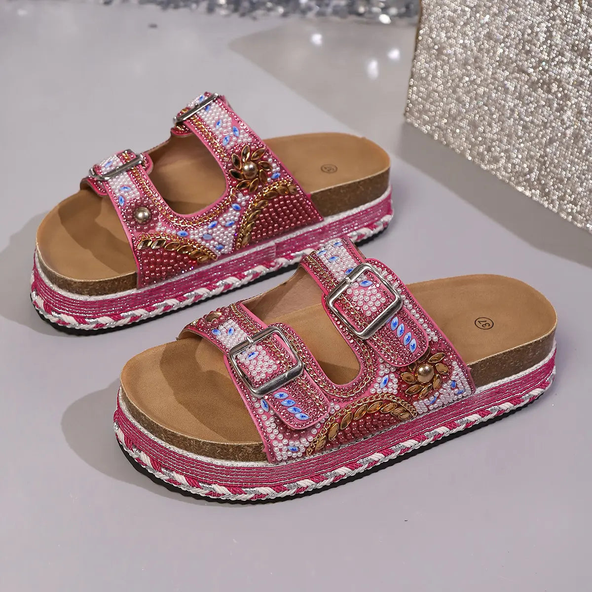 Pink platform slide sandals adorned with intricate bead and sequin embellishments, featuring double adjustable buckle straps and a cork-like sole with braided trim.