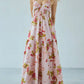 Bohemian floral gingham maxi dress on a model, featuring a smocked halter bodice, tiered flowing skirt, V-neckline, and cross-back straps, styled with tan ankle boots. The print combines pink gingham with red/pink flowers and green leaves.