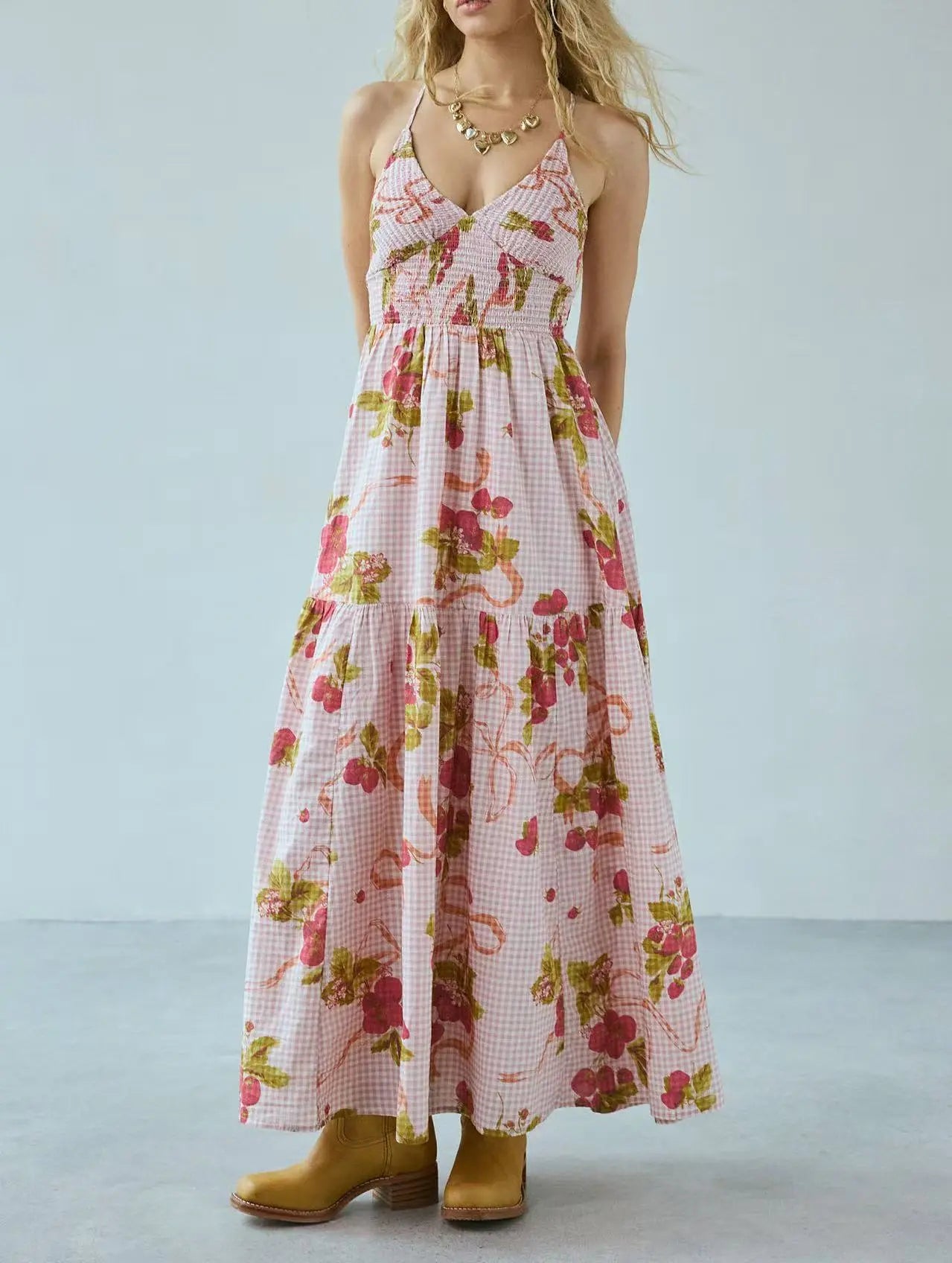 Bohemian floral gingham maxi dress on a model, featuring a smocked halter bodice, tiered flowing skirt, V-neckline, and cross-back straps, styled with tan ankle boots. The print combines pink gingham with red/pink flowers and green leaves.