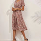 A woman models a pink ditsy floral print V-neck midi dress with short flutter sleeves and a cinched elasticated waist, paired with black high-heeled sandals. The dress falls to mid-calf.