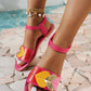 Women's bright pink flat sandals featuring playful multi-colored heart appliques in pink, yellow, red, and gold. Adorned with a black elasticated ankle strap, white contrast stitching, and a gold anklet, shown on a textured stone surface with a blurred blue water backdrop.