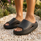 Kynso Obsidian Black platform lounge slides with thick, matte molded soles and side cutouts, worn by a person on a bright pebbled path amidst blurred greenery.