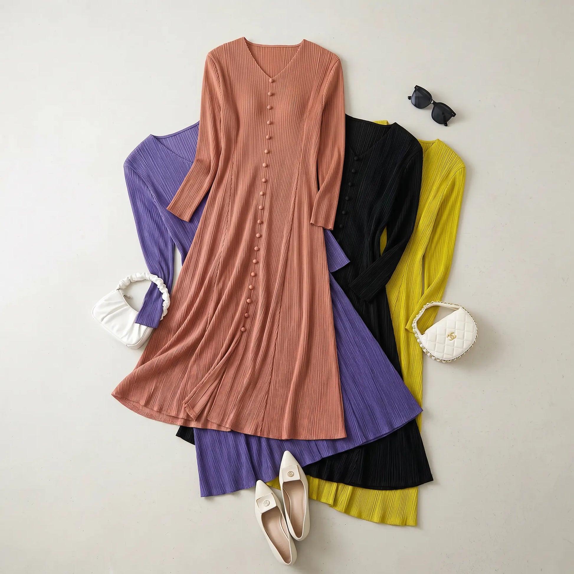 Flat-lay view of a rose pleated V-neck midi dress with fabric buttons, layered over purple, black, and yellow variants. Styled with white flats and handbags on a light background.