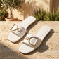 Pristine white square-toe flat slide sandals with wide vegan leather straps and prominent gold-tone D-ring buckles, styled outdoors on a textured stone.