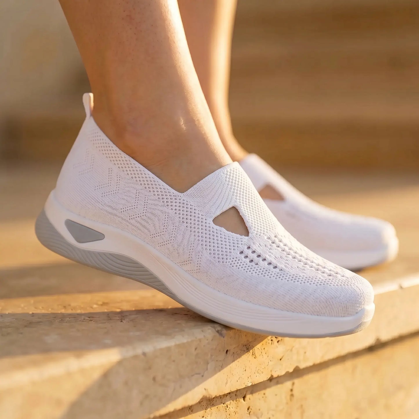 Pure White Ruxen slip-on sneakers on feet, showcasing the breathable woven knit upper with diamond perforations and a triangular instep cutout, alongside a grey heel accent. Worn on sunlit outdoor stone steps.