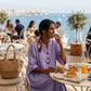 Smiling woman wearing a purple Mokari V-neck cotton linen midi dress with half sleeves and three buttons, enjoying brunch at a seaside cafe.