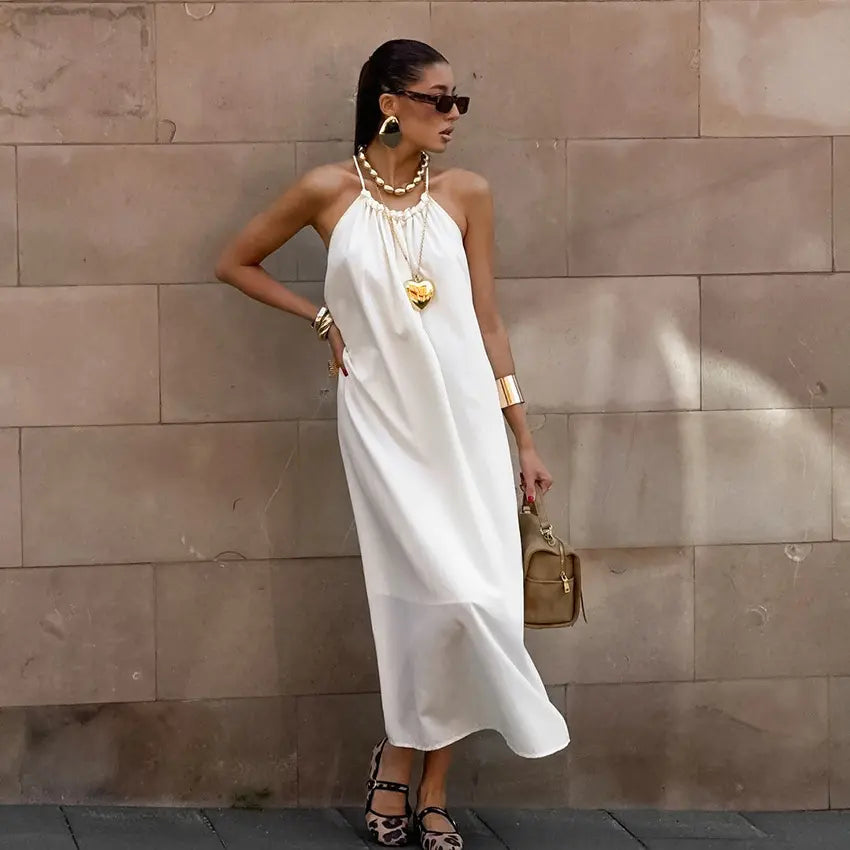 Model wearing Qyren's flowing ivory-white halter maxi dress with ruched neckline and A-line silhouette, styled elegantly against a textured stone wall.