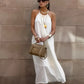 Model wearing an ivory white Qyren flowing halter maxi dress with a gathered neckline, elegantly draped against a neutral stone wall.