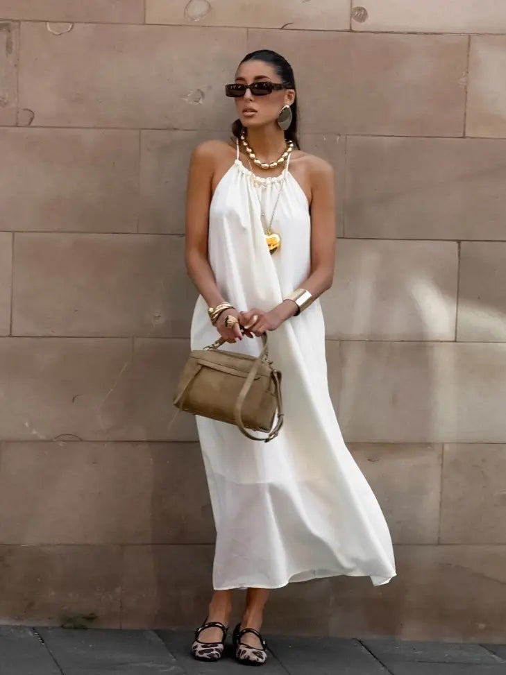 Model wearing an ivory white Qyren flowing halter maxi dress with a gathered neckline, elegantly draped against a neutral stone wall.