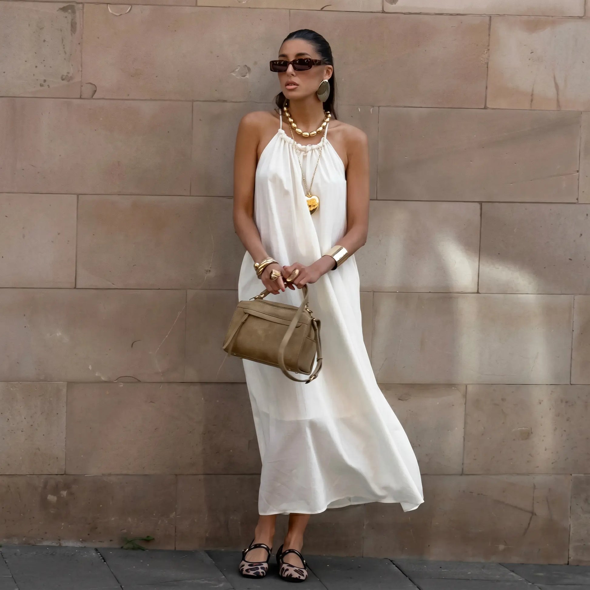 Model wearing an ivory white Qyren flowing halter maxi dress with a gathered neckline, elegantly draped against a neutral stone wall.