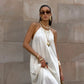 Model wearing a Qyren ivory-white flowing halter maxi dress with a ruched neckline, styled with gold jewelry against a stone wall.