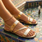 Radiant Gold Zyneth flat vegan leather sandals with distinctive asymmetrical cross straps and a comfortable elastic heel. Worn on feet on vibrant mosaic tiles.