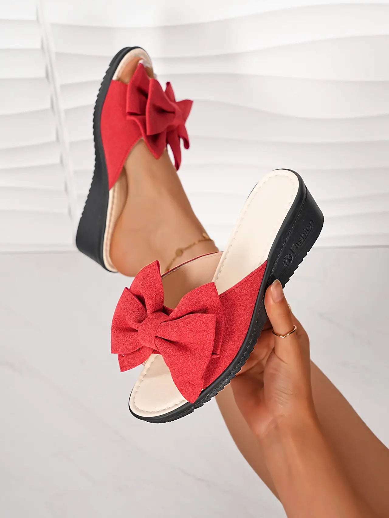 Red Lysa wedge slides featuring a large, soft bow detail on the single strap, shown on a foot with a black wedge heel and cream footbed against a studio background.