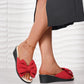 Red fabric wedge slides featuring a large, prominent bow detail on the strap, worn by a model with a dark skirt and anklet, showcasing a romantic and casual summer style.