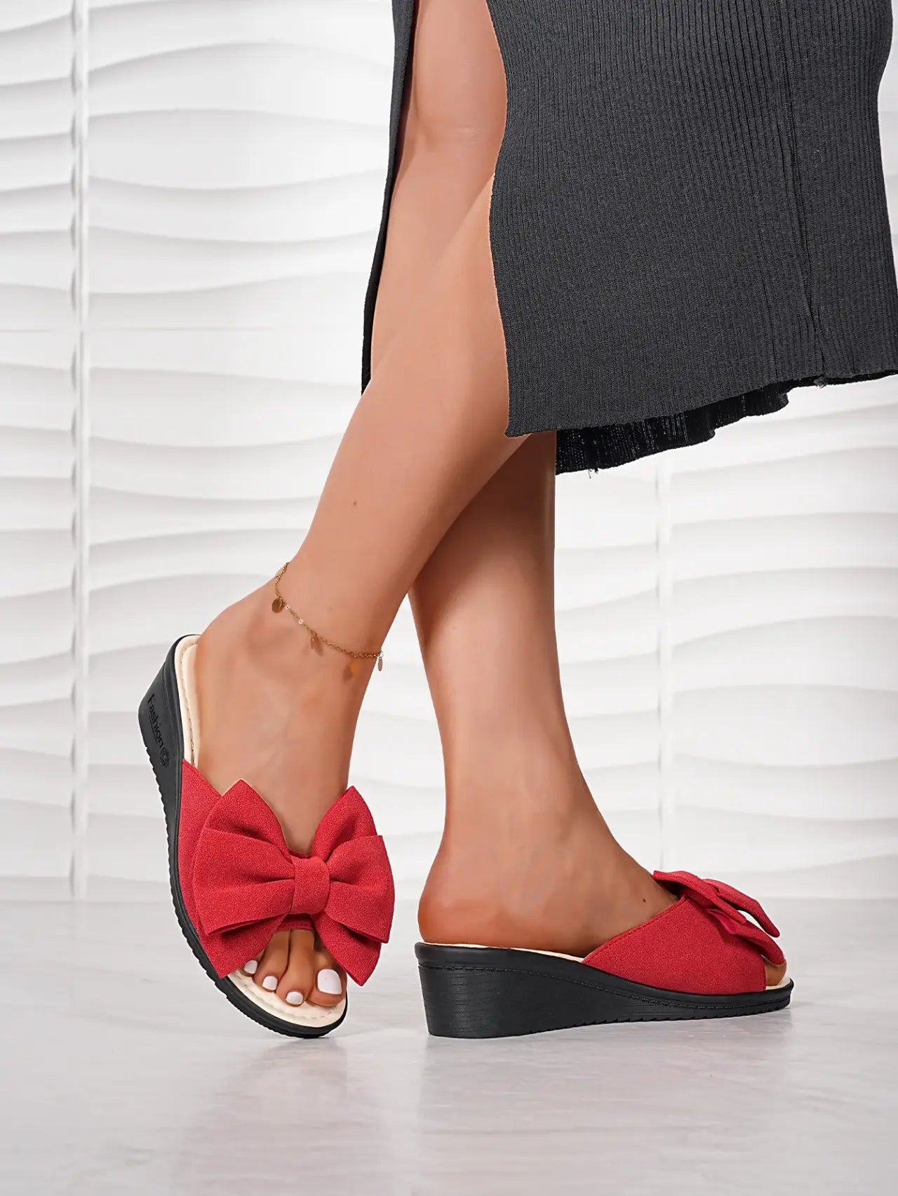 Red fabric wedge slides featuring a large, prominent bow detail on the strap, worn by a model with a dark skirt and anklet, showcasing a romantic and casual summer style.