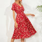 Red midi dress with a ditsy floral print, featuring a V-neck, short flutter sleeves, and a cinched smocked waist, shown on a model against a textured background with palm leaf shadows.
