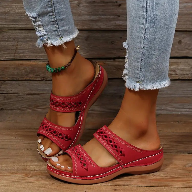 A pair of women's red slip-on wedge sandals with geometric embroidered straps, worn with distressed denim jeans and a beaded anklet.