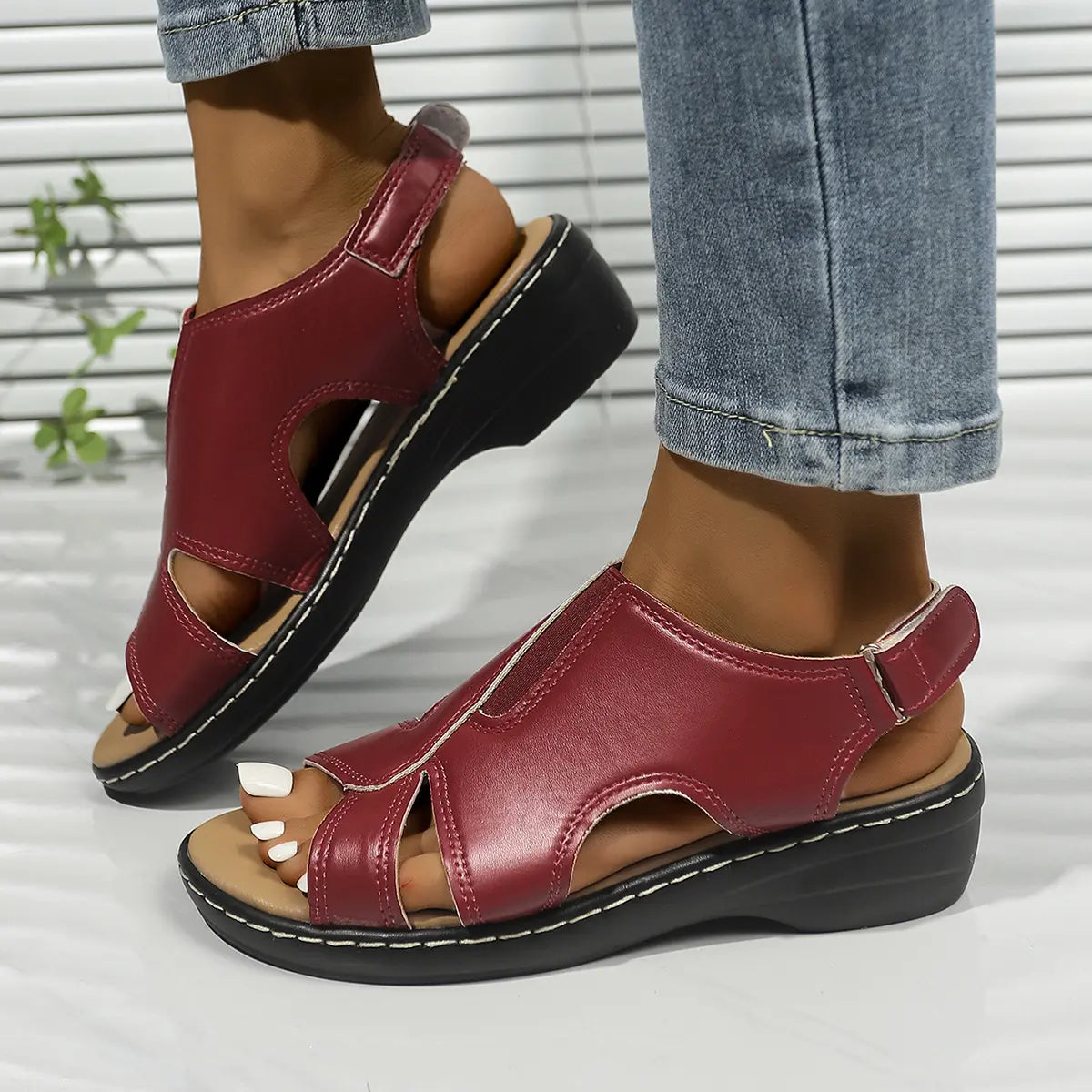 Red faux leather peep-toe wedge sandals with an adjustable ankle strap and white contrast stitching, shown on a model's feet.
