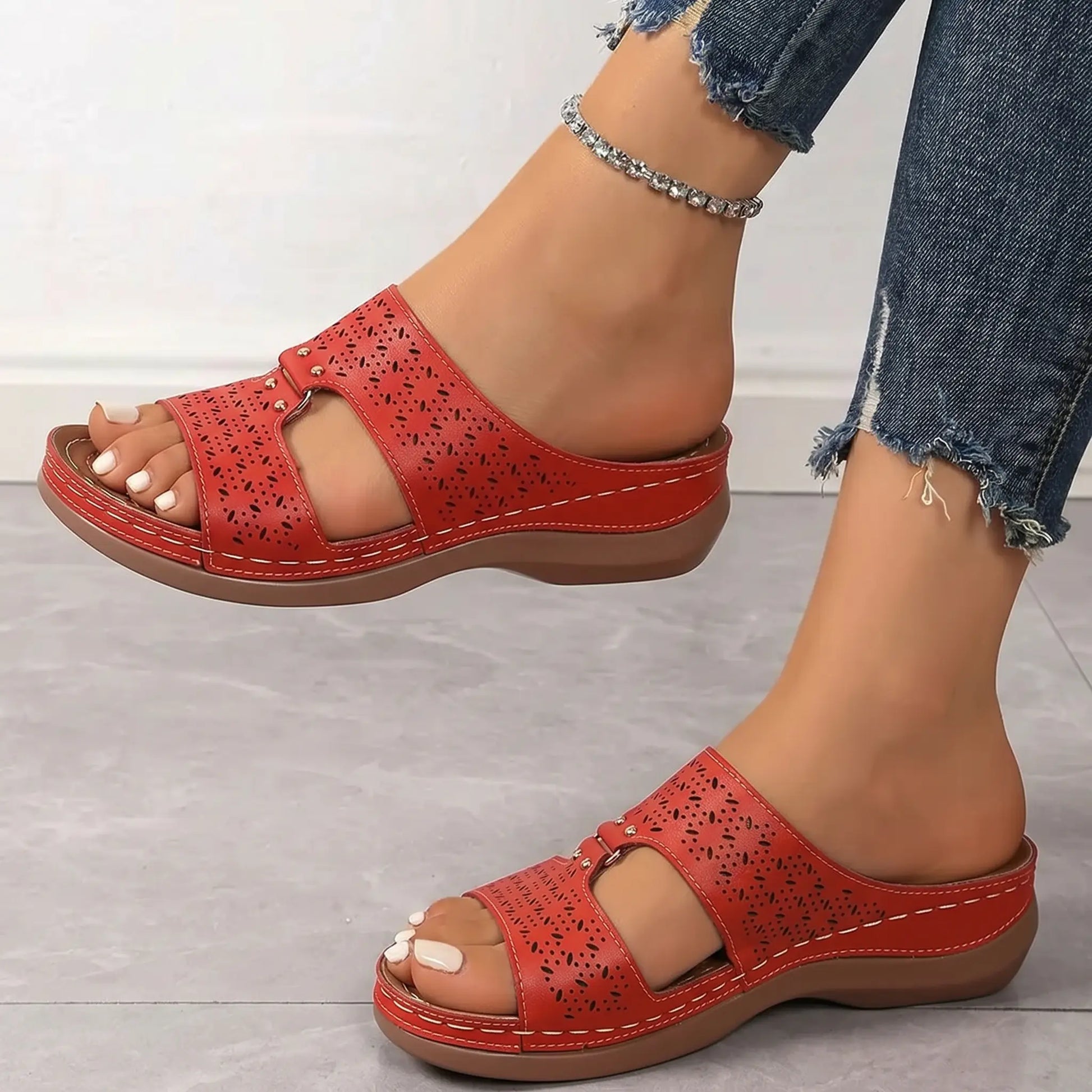 Side view of a model's foot wearing vibrant red Briska slip-on sandals featuring intricate laser-cut patterns, metallic stud accents, and a low wedge heel, paired with ripped blue jeans.
