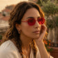 A woman with curly brown hair wears vibrant crimson octagonal sunglasses with thin gold-tone metal frames, set against a sun-drenched outdoor backdrop with blurred buildings and foliage.