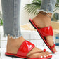 Red patent toe-thong flat sandals with a braided toe loop worn with distressed denim jeans and a gold anklet, showcasing the glossy finish and comfortable slip-on style.